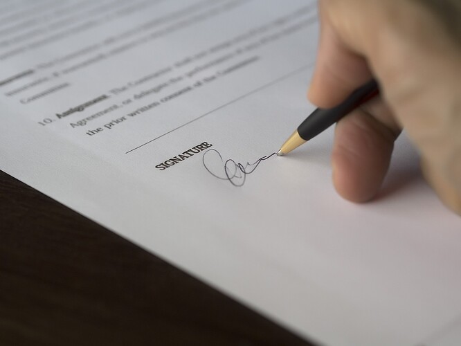 A hand holding a pen, signing a contract of work. The pen is touching the paper under the heading of "Signature", with a partial signature drawn